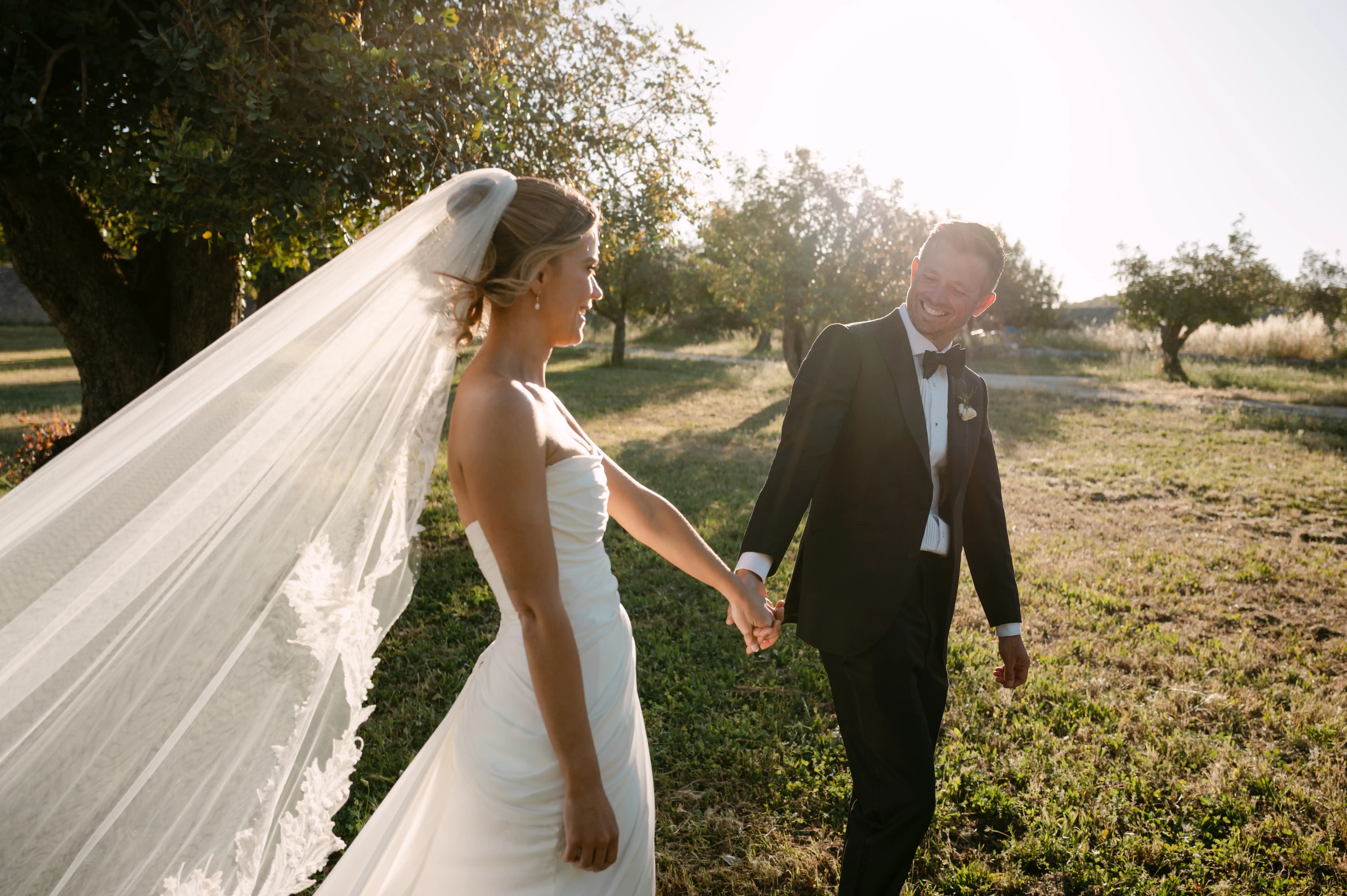 Bride and groom holding hands while walking through golden sunset light at a countryside wedding in Italy.
